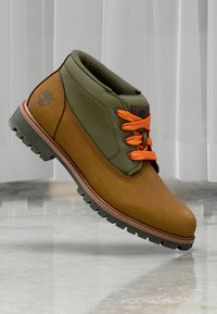 Botte de randonnée marron et verte en cuir et textilé, avec des lacets orange, une semelle en caoutchouc texturée et un logo sur le côté.