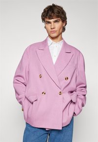 Rosa dobbeltspent blazer med gullknapper, teksturert stoff, klaffelapper og sidelommer, parret med en hvit skjorte og blå jeans.