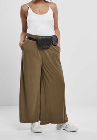 Femme portant un débardeur blanc, un pantalon large olive, des baskets blanches et un sac banane en cuir noir, debout les mains dans les poches.