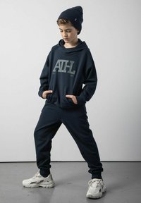Granatowy hoodie z dużym szarym logo, w połączeniu z dopasowanymi dresami. Uzupełniony o wełnianą czapkę i białe buty sportowe.