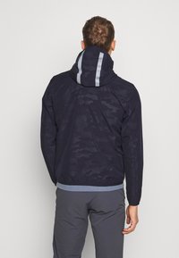 Marineblå hættejakke med camouflage mønster, kontrasterende lysegrå striber på hætten og en ribbet blå kant.