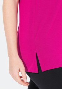 Top fucsia hecho de una tela suave, con una abertura lateral y un corte relajado, con leggings negros parcialmente visibles en el fondo.