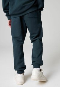 Tamsiai žali treningai, pagaminti iš minkštos medžiagos, su šoninėmis kišenėmis ir užrišamais kraštais, derinami su baltais sportbačiais su šviesiais akcentais.