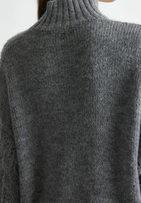 Helles graues, übergroßes gestricktes Sweatshirt mit hohem Rippkragen, das auf der Rückseite ein kleines Logodetail aufweist. Die Textur wirkt weich und gemütlich.
