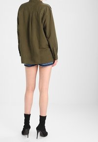 Camicia oversize verde oliva, tessuto morbido al tatto, dettagli con bottoni, maniche lunghe. Indossata sopra pantaloni denim e stivali neri alla caviglia.