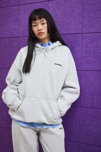Giovane donna con lunghi capelli neri che indossa un felpa grigia chiara e pantaloni, in piedi con le mani nelle tasche contro un muro di piastrelle viola.
