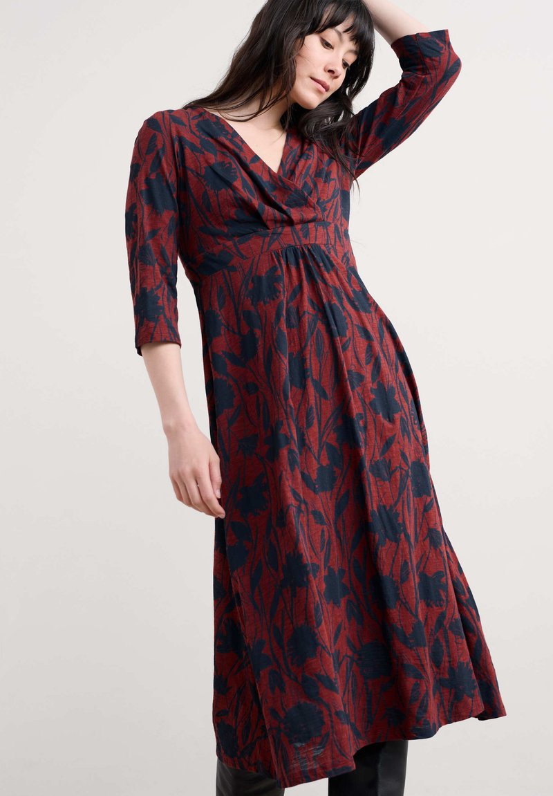 Femeie îmbrăcată într-o rochie midi cu mânecă lungă, cu un model floral roșu și negru, pozând cu un braț ridicat și capul aplecat în jos.