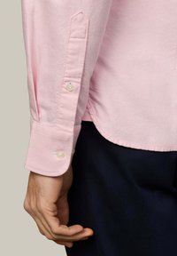 Ľavá ruka v svetloružovej košeli s dlhým rukávom a zapnutým manžetovým gombíkom, pod lemom košele sú viditeľné tmavé nohavice.