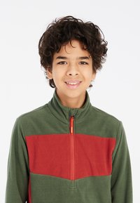 Sudadera de forro polar en verde con una franja horizontal roja, que cuenta con un collar alto y una cremallera frontal con un tirador naranja.