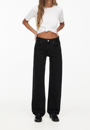 Jean large noir avec taille haute et cinq poches, associé à un t-shirt gris ajusté noué à la taille. Texture en denim lisse.