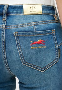 Κοντινό πλάνο της πίσω τσέπης ενός μπλε τζιν με κεντημένο άλογο σε κόκκινο και κίτρινο χρώμα και ετικέτα "Armani Exchange".