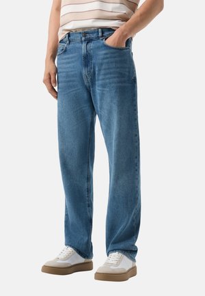 Mann trägt blaue Jeans mit geradem Bein und weiße Sneaker, Hand in der Tasche, vor einfarbigem Hintergrund.