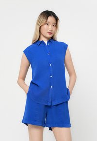 Blaues ärmelloses Hemd mit Knopfleiste, runder Saum und zwei seitlichen Taschen; kombiniert mit passenden blauen Shorts. Glatte Textur.
