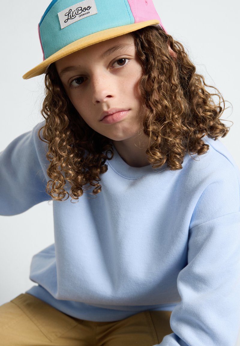 Hellblaues Sweatshirt mit Rundhalsausschnitt; beige Hose. Buntes Cap mit türkisfarbenen, pinken und gelben Paneelen sowie einem Logo-Patch. Lockige Haare.