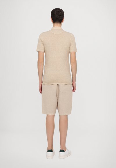 Polo de manga corta beige con una textura suave, combinado con pantalones cortos beige claros. Zapatillas blancas con acentos verdes completan el conjunto.