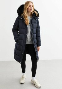 Mörkblå dunjacka med fuskpäls på huvan, tryckknapp och dragkedjestängning, quiltat mönster, kombinerad med svarta leggings och beige sneakers.
