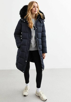 Manteau matelassé navy avec une capuche en fausse fourrure, fermeture à boutons-pression et à glissière, design matelassé, associé à des leggings noirs et des baskets beiges.