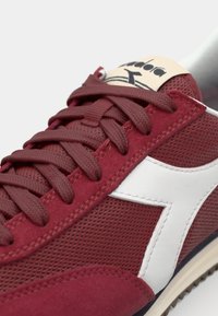 Red suede and mesh sneakers feature white accents, textured laces, and a branded tongue. The sole has a contrasting beige rubber tread.