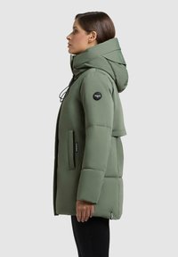 Giacca a vento verde con colletto alto, tasche frontali e cappuccio regolabile. Presenta una texture liscia e una patch con logo sulla manica.