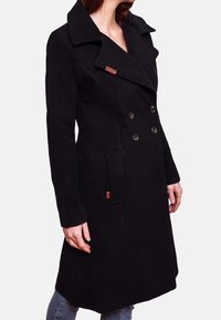 Donna con un cappotto nero doppiopetto con ampi rever e tasche anteriori, abbinato a jeans blu, su uno sfondo semplice.