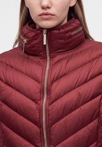 Femme portant une veste matelassée bordeaux à col montant avec des fermetures éclair dorées sur le devant et le col, affichée de la poitrine à la bouche.