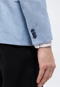 Χέρι άνδρα δίπλα σε μανίκι ανοιχτό μπλε σακακιού με τρία σκούρα κουμπιά, ορατή μανσέτα λευκού πουκάμισου και μαύρο παντελόνι.