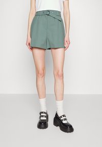 Pantaloni corti verdi su misura con vita con cintura, dotati di tasche anteriori e un design pulito e strutturato. Indossati con scarpe nere chunky e calzini bianchi.