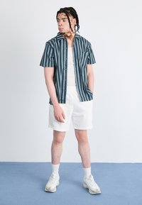 Striped short-sleeve shirt in dark teal and white, paired with white shorts and light sneakers. Features a button-up front and chest pocket.