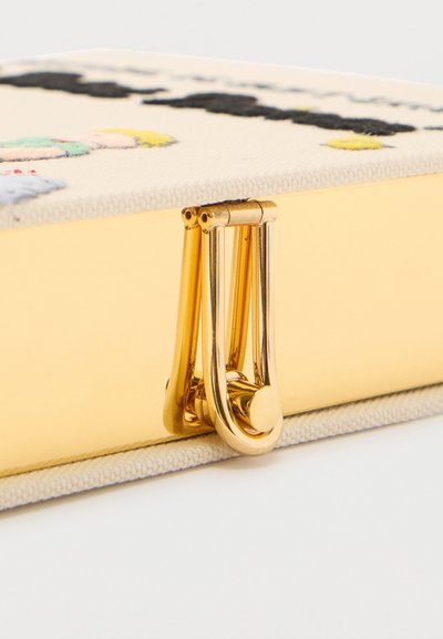 Broche de oro en el borde de un objeto rectangular con tela ligera y detalles bordados coloridos en la parte superior, sobre un fondo blanco.