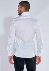 Mann mit kurzen Haaren und Bart, der ein tailliertes weißes Langarmshirt und schwarze Hosen trägt und von einem schlichten Hintergrund abgewandt ist.