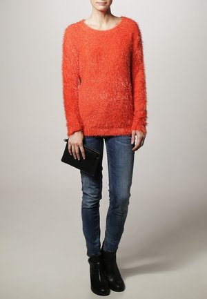 Un pull orange duveteux avec un col rond, associé à un jean skinny bleu et des bottes noires à la cheville. Une petite pochette noire est tenue dans une main.