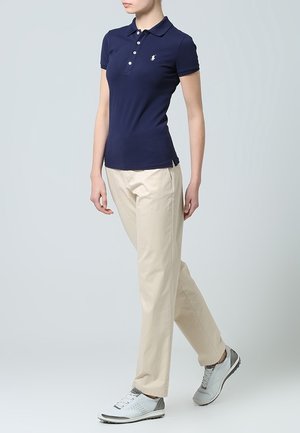 Kvinna klädd i en marinblå piké, beige byxor och vita sneakers, stående med ena benet något korsat framför det andra.