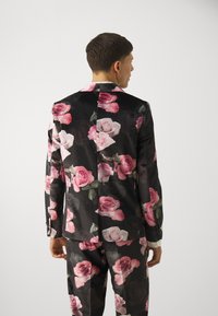 Black velvet suit with large pink and white rose patterns. Designed with a notch lapel and tailored fit, featuring buttoned cuffs and striking floral accents.