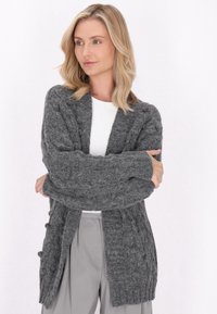 Cardigan gri tricotat cu modele în cablu, cu deschidere frontala, mâneci lungi și borduri cu nervuri. Textură moale și design până la genunchi.