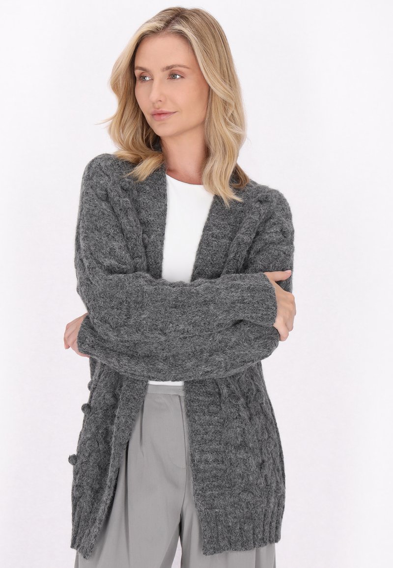 Cardigan gri tricotat cu modele în cablu, cu deschidere frontala, mâneci lungi și borduri cu nervuri. Textură moale și design până la genunchi.