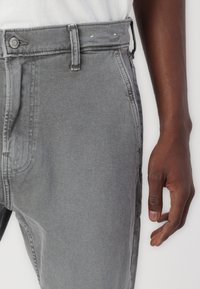 Pantalones vaqueros de mezclilla gris con textura lisa, corte de tiro medio, trabillas para cinturón y costuras en contraste. Cierre frontal con botón y cremallera.