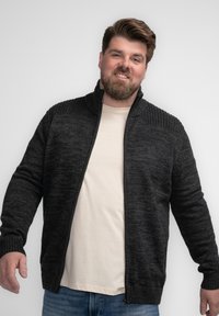Cardigan oscuro gris con cremallera y cuello texturizado, puños acanalados y patrón de punto vertical. Lleva puesto encima de una camiseta de color claro.