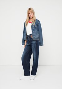 Denim striukė, mėlyna, su sagomis, su dviem priekiniams kišenėm; derinama su aukštu liemeniu plačiomis mėlynomis džinsinėmis kelnėmis ir baltais sportbačiais.