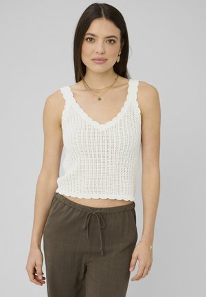 Femme portant un haut sans manches en crochet blanc à bords festonnés et un pantalon olive à cordon de serrage, sur fond gris clair