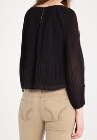 Kvinna som bär en svart långärmad plisserad blus med kallaxelutskärningar och beige byxor med sydda bakfickor.