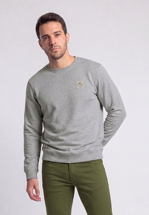 Homme portant un sweat-shirt gris clair et un pantalon vert olive debout devant un fond clair uni.