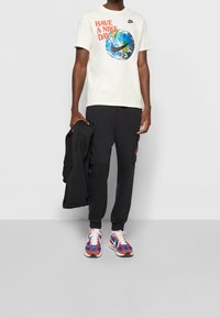 Nike T-paita vaaleassa kremessä, jossa on maapallon grafiikka ja punainen teksti, yhdistettynä mustiin cargo-housuihin ja monivärisiin tennareihin, joissa on punaisia, sinisiä ja valkoisia yksityiskohtia.