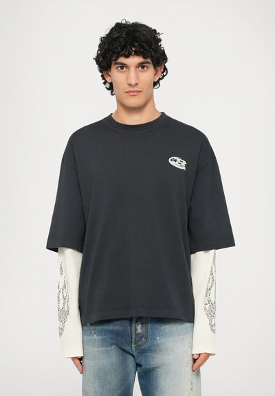 T-shirt nero oversize con colletto rotondo, presenta una grafica bianca sul petto. Sovrapposto a una maglietta a maniche lunghe bianca con un motivo grigio.