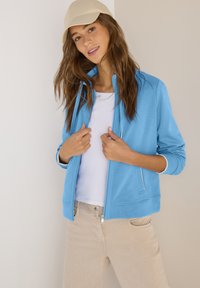 Femme aux longs cheveux bruns portant une casquette beige, une veste zippée bleu clair sur un t-shirt blanc, et un pantalon beige, posant devant un fond uni.