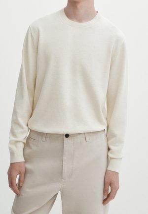 Persoon die een crèmekleurige trui met lange mouwen draagt, ingestopt in lichtbeige broek met een zwarte knoop en zijzakken.