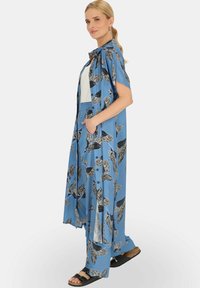 Niebieski długi kardigan z krótkimi rękawami, zdobiony czarnymi wzorami liści, z bocznymi kieszeniami i luźnym krojem, zestawiony z białym topem i sandałami.