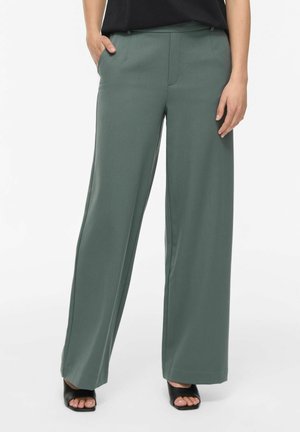Femme portant un pantalon vert taille haute à jambes larges et des talons ouverts noirs, une main dans la poche et un haut noir.