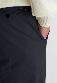Persoon met donkerblauwe broek met achterzak met knoop en hand in zijzak, gecombineerd met een crèmekleurige, geribbelde sweatermouw.