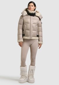 Beige Daunenjacke mit abnehmbarem, gefälschtem Futterkapuze, schwarzen Akzenten und Seitentaschen. Tragen mit hautfarbenen Leggings und weißen Stiefeln.