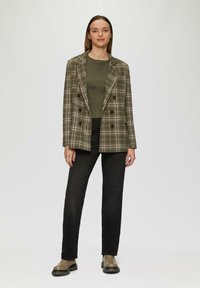 Doppelreihiger, karierter Blazer in Grün und Beige über einem figurnahen oliven Oberteil, kombiniert mit schwarzen, geraden Jeans und oliven Stiefeletten.